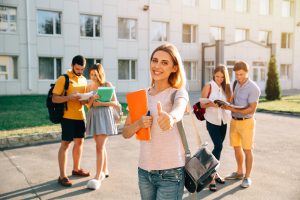 Happy beautiful girl standing with note books and backpack showing thumb up and smiling, standing near university building, her friends are behind